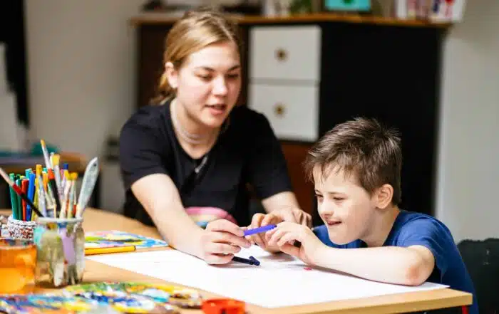 Close up of young female teacher sitting at desk with a Down syndrome schoolboy. Color painting on the paper for disabled kids, autism childs who are down syndrome and student teacher.