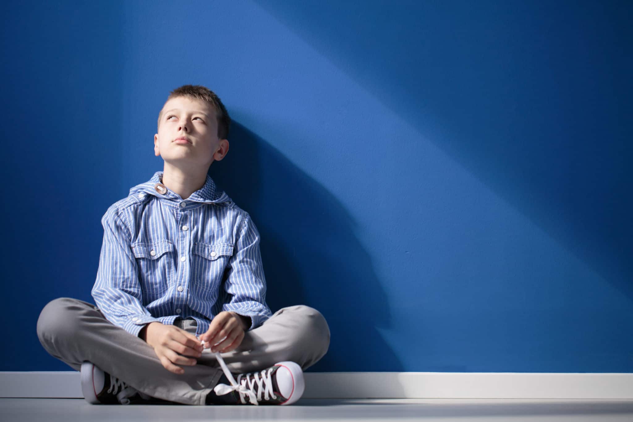 Thoughtful autistic boy sits with crossed legs on white floor against blue wall