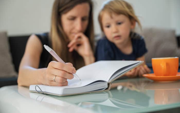 Young student mother studying from home, making notes in notebook with her toddler daughter next to her.
