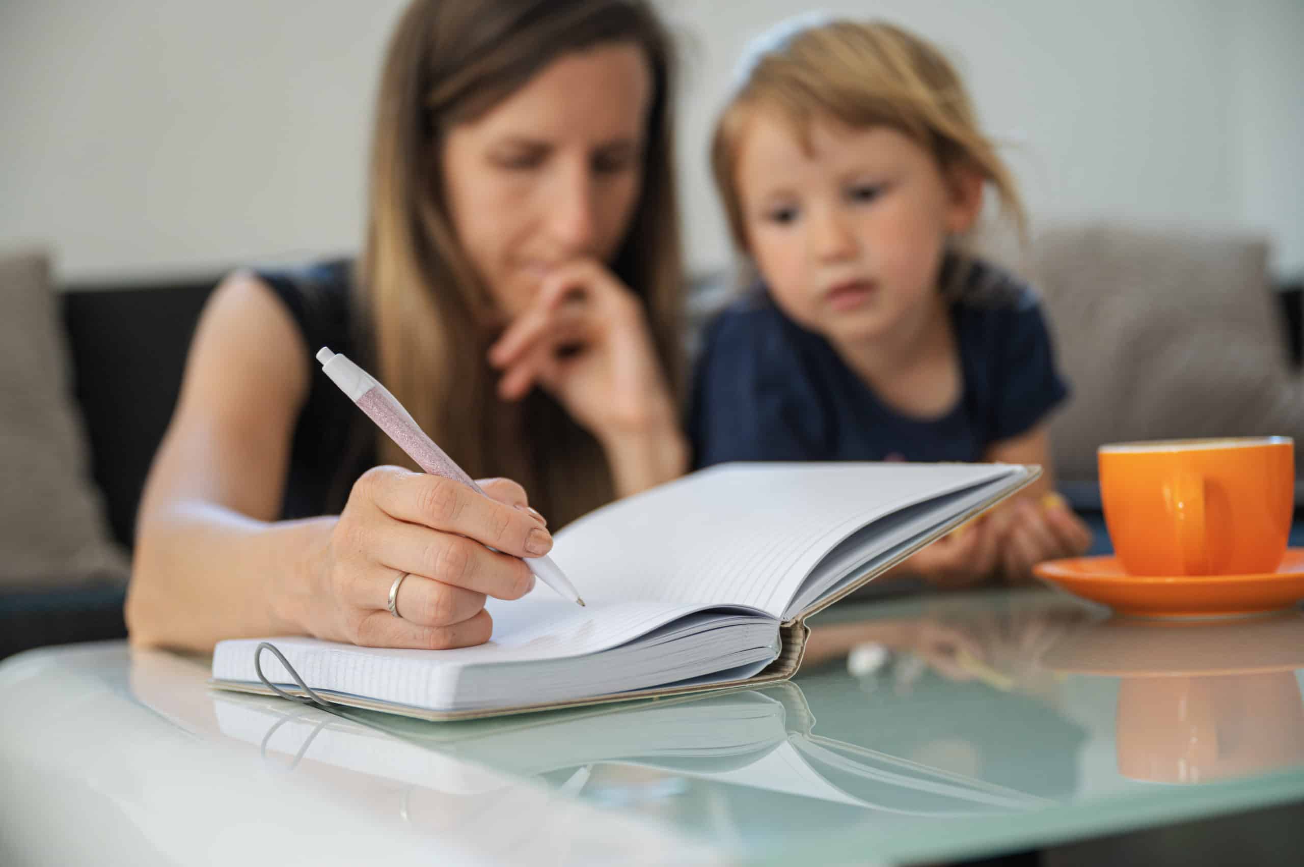 Mother writing in notebook with child Young student mother studying from home, making notes in notebook with her toddler daughter next to her.