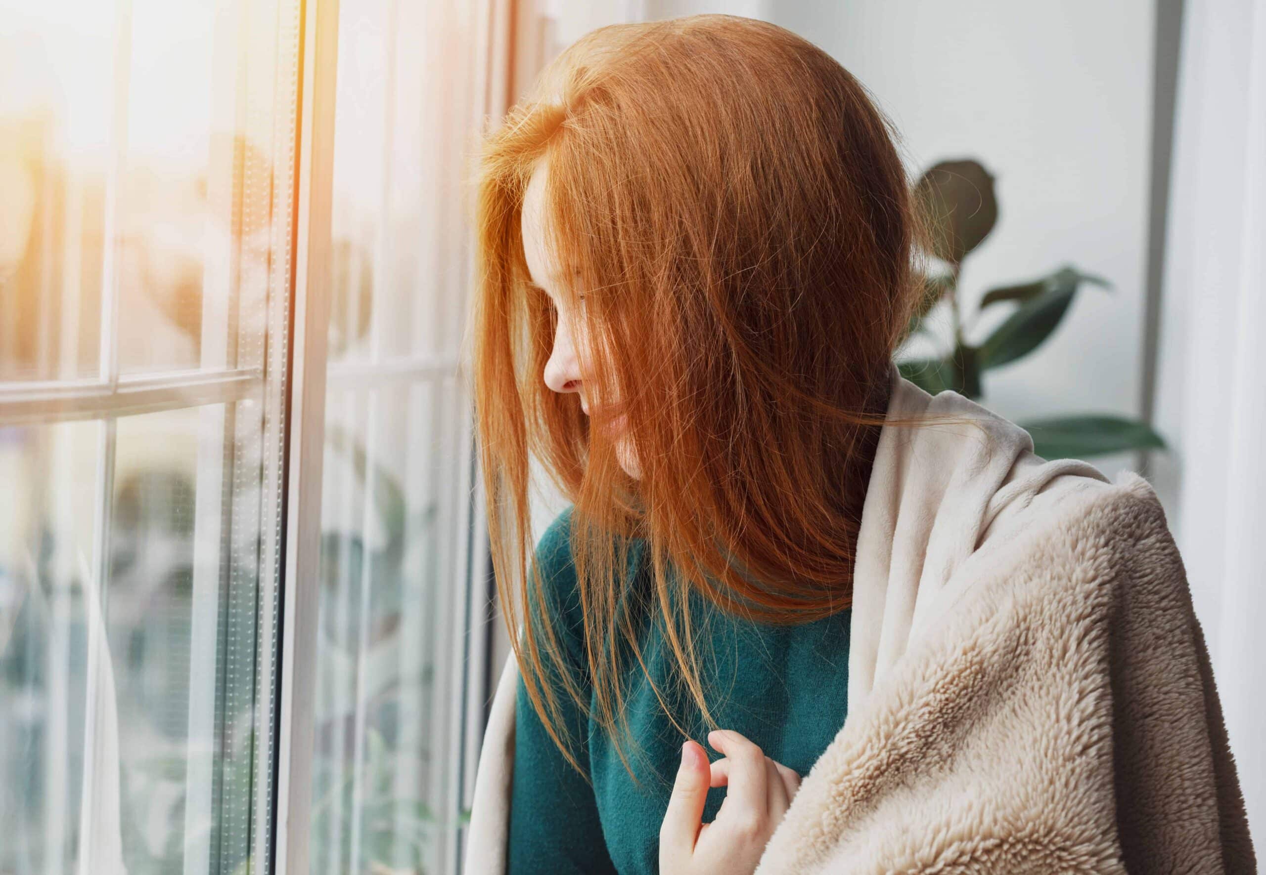 beautiful thoughtful pensive calm smiling young woman with long red hair looking through the window at home wrapped in warm comfy blanket Close-up of a woman with ginger hair, looking outside through a window