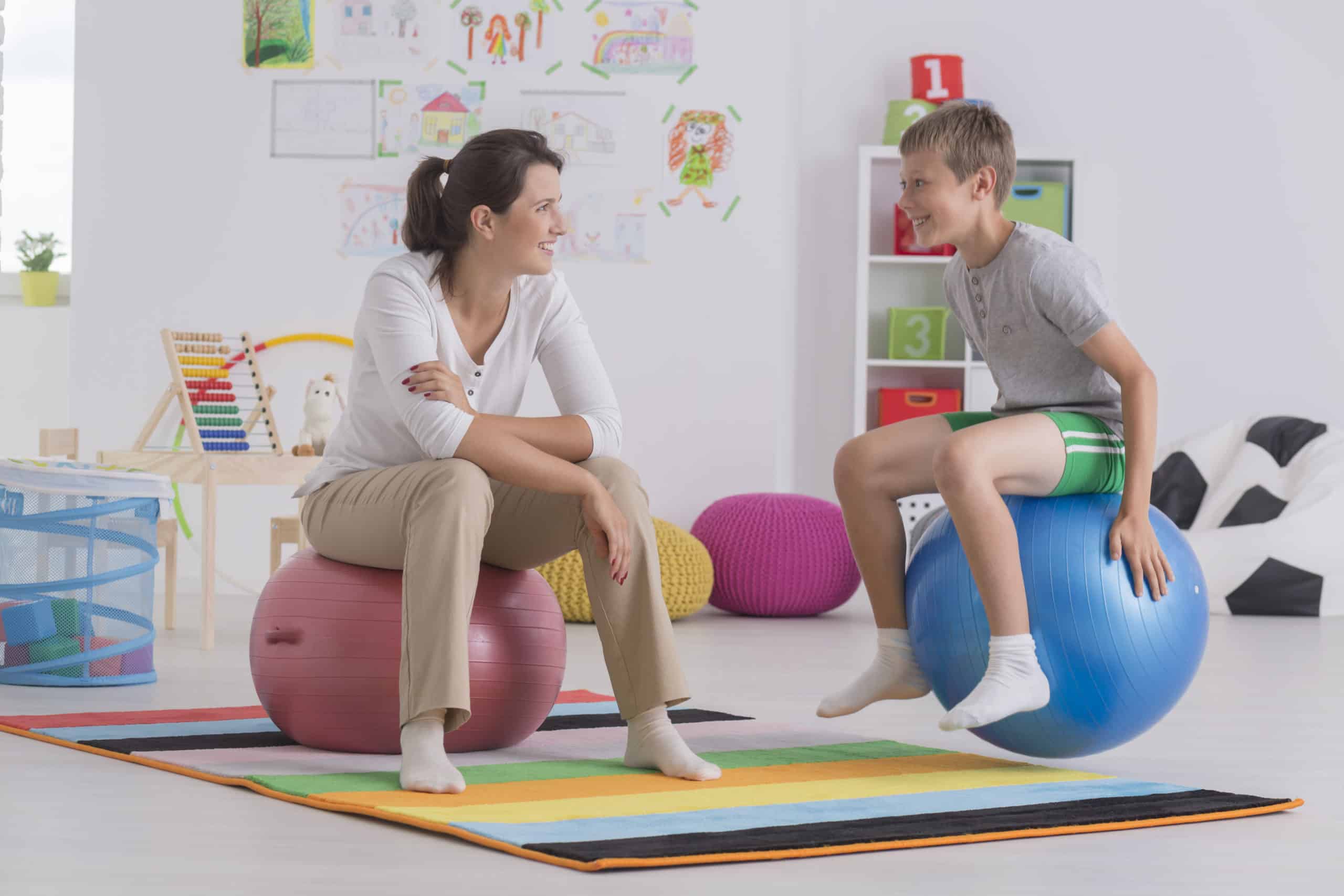 Physiotherapist and happy boy sitting on gym ball