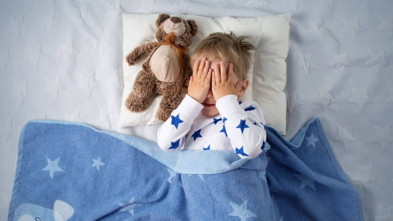 Kid resting on a bed with a teddy bear beside him, covering his face