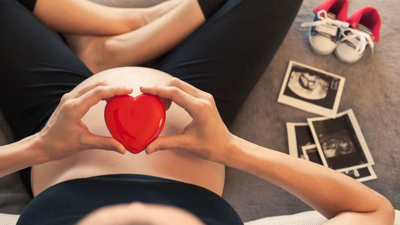 Pregnant woman holding a red heart-shaped figure with baby shoes beside her