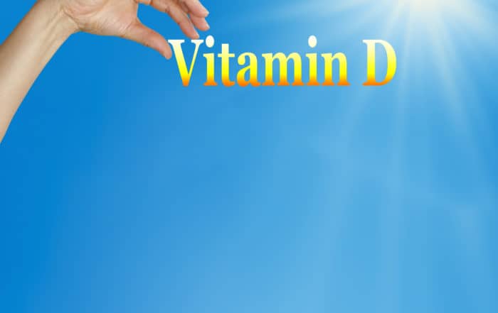 Hand holding up the words "vitamin D" on blue sky background with sunshine coming from upper right.