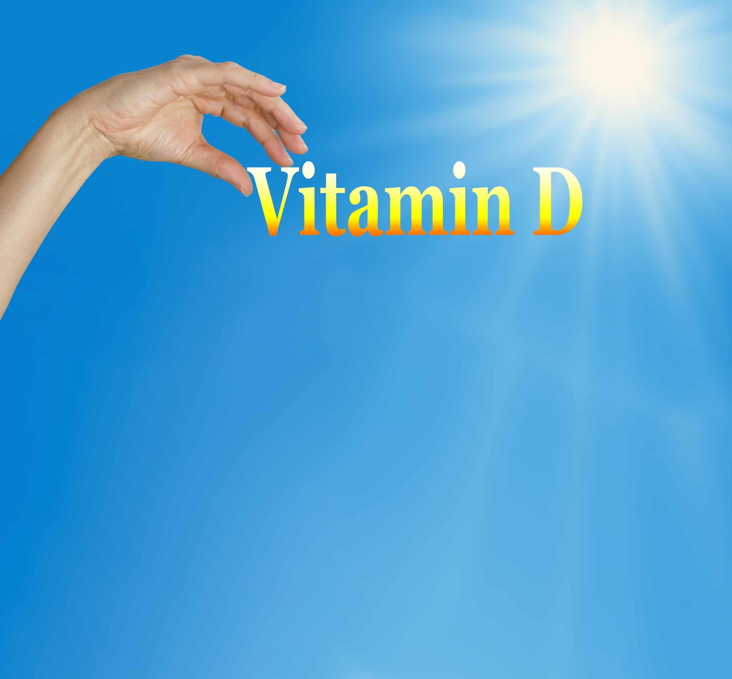 vit d Hand holding up the words "vitamin D" on blue sky background with sunshine coming from upper right.